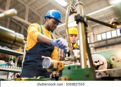 Multi-ethnic team of workers wearing overalls and protective helmets using lathe in order to machine workpiece, interior of spacious production department on background