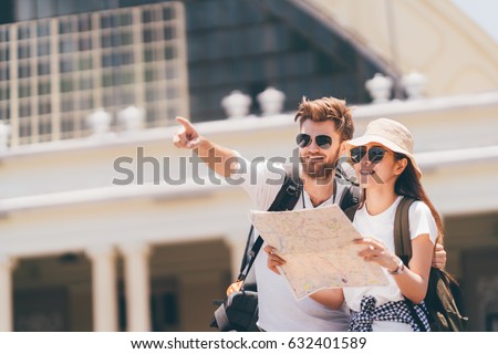 Multi-ethnic traveler couple using local map together on sunny day, man pointing forward to copy space. Honeymoon trip, backpacker tourist, Asia city tourism, or summer holiday vacation travel concept