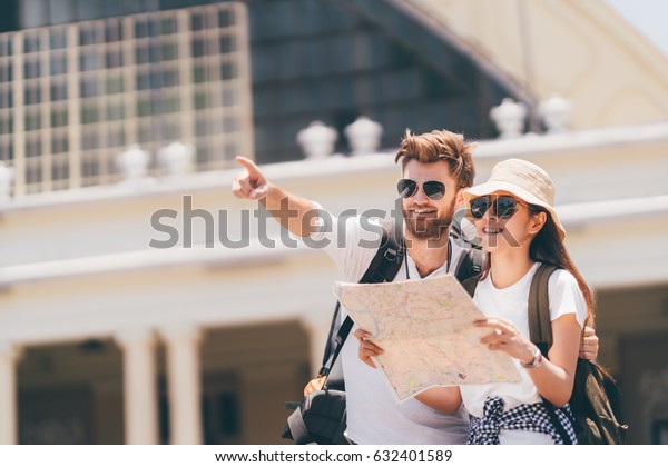 Multi-ethnic traveler couple using local map\
together on sunny day, man pointing forward to copy space.\
Honeymoon trip, backpacker tourist, Asia city tourism, or summer\
holiday vacation travel\
concept