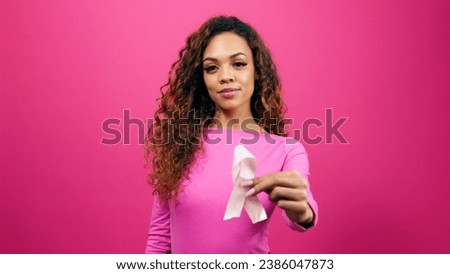 Multi-ethnic woman holds breast cancer awareness ribbon to camera, pink studio