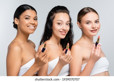 multiethnic women with bare shoulders smiling while holding lipsticks isolated on grey
