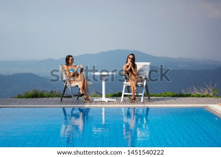 Multi-ethnic women in swimsuits drinking cocktails by swimming pool with mountain view on summer holidays