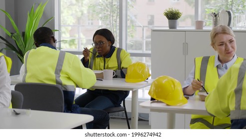 Multiethnic Workers In Safety Uniform Eating Meal In Factory Canteen. Construction Company Engineers And Architects Sitting At Table And Having Lunch Break