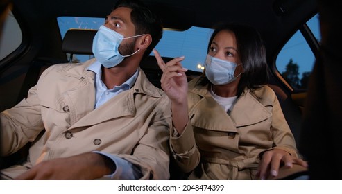 Multiethnic Young Couple In Safety Mask Sitting On Backseat In Car. Diverse Colleagues Wearing Protective Mask Sitting In Taxi Commuting Together