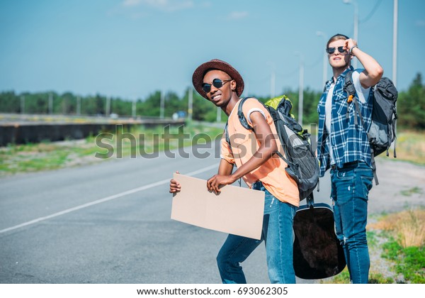 multiethnic young men with empty cardboard\
looking for car while hitchhiking during\
trip