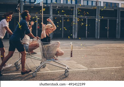 Multiethnic young people racing with shopping cart and blowing confetti. Young friends having fun on shopping carts. - Shutterstock ID 491412199