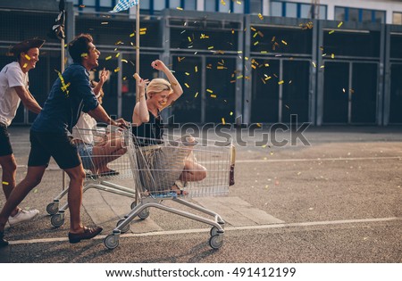 Multiethnic young people racing with shopping cart and blowing confetti. Young friends having fun on shopping carts.