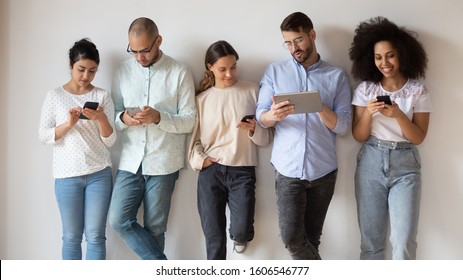 Multiethnic Young People Stand In Row Near White Wall Busy Using Electronic Devices Browsing Wireless Internet, Diverse Millennial International Employees Obsessed Addicted To Gadgets And Technology