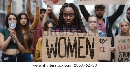 Multi-ethnic young protesters marching for women rights, right to abortion, showing a signboard of Women. African woman in front holding the sign about woman empowerment.