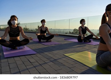 Multiethnic Young Women Group Wear Face Masks Meditate In Lotus Pose Keep Social Distance At Retreat Yoga Class On Sunrise Outdoor. Fit Healthy Diverse Ladies Do Yoga Exercise Distancing For Safety.