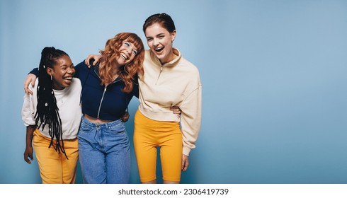 Multiethnic young women laughing and having fun while embracing each other. Group of cheerful female friends enjoying themselves while standing against a blue background.