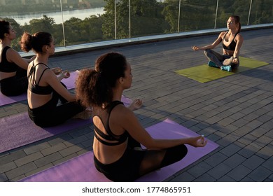 Multiethnic young women meditate with yoga teacher in lotus pose breath fresh air at retreat yoga class on sunrise outdoor.Fit healthy multiracial ladies do yoga exercise in summer sunset together.