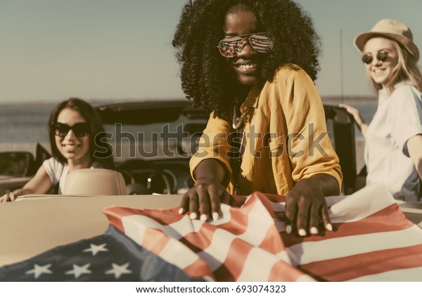 multiethnic young women resting in car with\
american flag at\
seaside