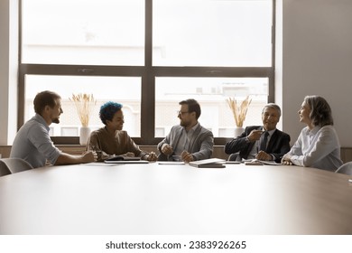 Multiethnic younger and elder business coworkers sitting in row together at large meeting table, talking, laughing, enjoying informal conversation, communication, networking - Powered by Shutterstock