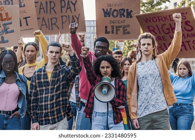 Multiethnic Youth Protesting for Decent Work and Life - A group of multiethnic youth protesting for fair pay and work access, raising fists. A curly-haired woman shouts slogans through a megaphone