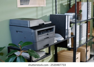 Multifunction printer standing on office shelf surrounded by green plant, office supplies, and shelves with boxes and folders creating organized workspace