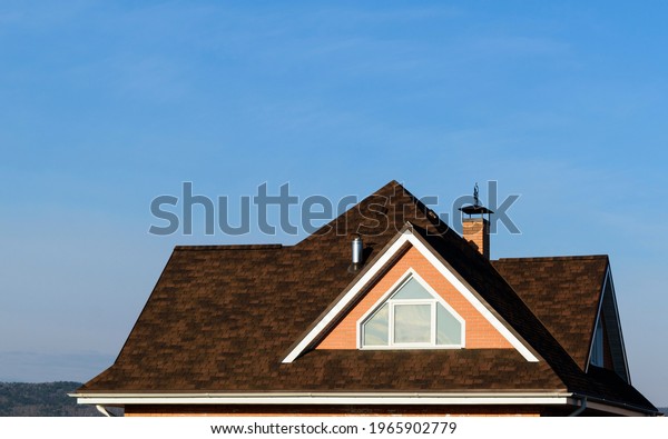 Multi-gable roof of house. Roof window. Flexible\
roofing material in brown color. Brick, chimney pipes. Background -\
blue sky.