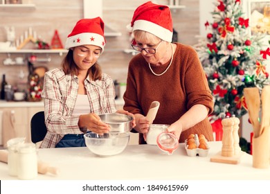 Multigeneration family doing cookies together on christmas day. Happy cheerful joyfull teenage girl helping senior woman preparing sweet buiscuits to celebrate winter holidays wearing santa hat.