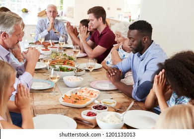 Multi-Generation Family And Friends Around Table Praying Before Meal At Party