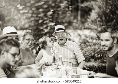 Multi-generation Family Gathered To Lunch In The Garden In Summer, They Having Fun Sitting Around A Picnic Table. Black And White