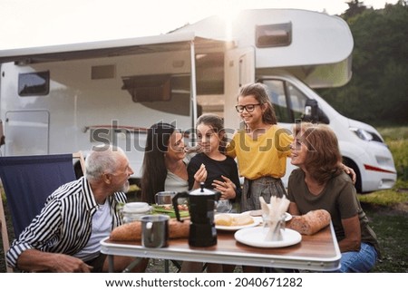 Multi-generation family sitting and eating outdoors by car, caravan holiday trip.