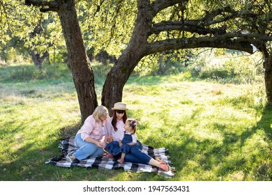 Multi-generation family spending time outdoors in sunny summer garden, sitting on checkered blanket under the big apple tree. Mature grandmother with daughter and granddaughter in park. - Shutterstock ID 1947563431