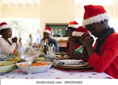 Multigeneration Family Wearing Santa Hats Praying Before Having Lunch Together While Sitting On Dining Table In The Living Room At Home. Christmas Festivity Tradition Celebration Concept