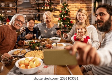 Multigenerational Caucasian family taking a selfie on smartphone while eating dinner at home during Christmas and the new year holidays - Powered by Shutterstock