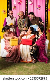 Multigenerational Indian Family Eating Sweets While Celebrating Festival Or Occasion Dressed In Traditional Wear, Sitting On Sofa/couch