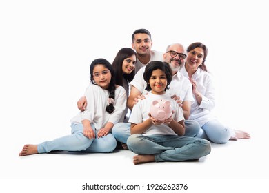 Multigenerational Indian Family Of Six Holding Piggy Bank While Wearing White Cloths And Standing Against White Wall