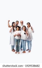 Multigenerational Indian Family Of Six Holding Piggy Bank While Wearing White Cloths And Standing Against White Wall
