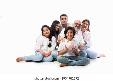 Multigenerational Indian Family Of Six Holding Piggy Bank While Wearing White Cloths And Standing Against White Wall