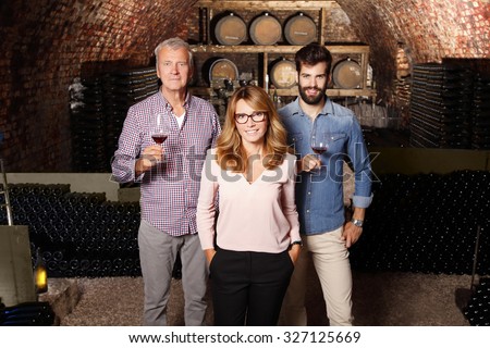 Multigenerational winery owner family standing at wine cellar. Senior winemaker and young sommelier holding a glass of red wine while businesswoman looking at camera and smiling. Small business. 