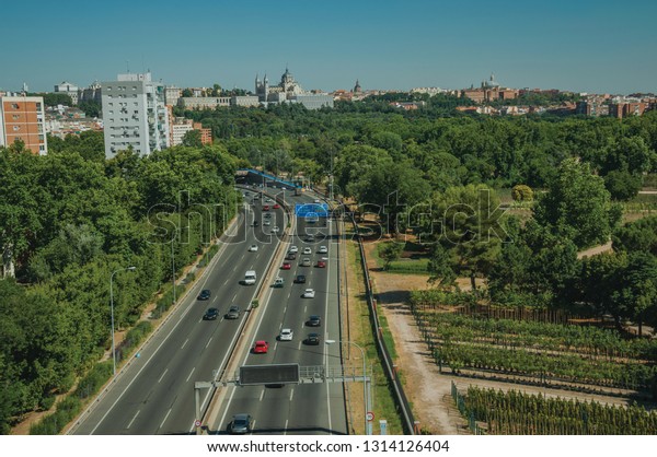 Multi-lane highway with heavy traffic in the midst\
of garden trees and apartment buildings, in a sunny day at Madrid.\
Capital of Spain this charming metropolis has vibrant and intense\
cultural life.