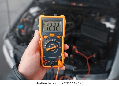 Multimeter in hand, checking car battery. Check car battery using voltmeter. Man check up voltage level, alternator produce 12.3 volts with the engine idling. Winter service maintenance
