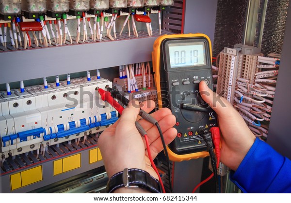 Multimeter is in hands of electrician on\
background of electrical automation cabinet. Adjustment of\
electrical control circuit for industrial\
equipment.