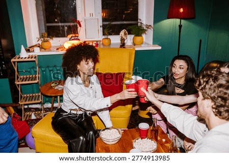 A multinational group of young people making a toast with red cups in a colorful, cozy living room decorated for a birthday party, surrounded by snacks, popcorn bowls, and warm lighting.