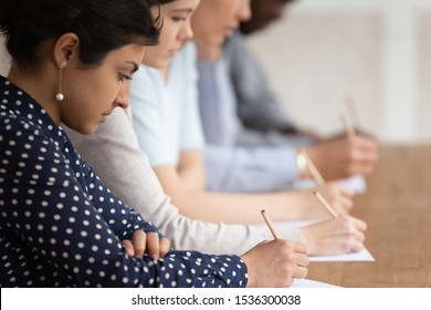 Multinational students seated at desk in row holding pencils writing on papers, take part in university exams, learning process or test of scholars knowledge skill in subject, higher education concept
