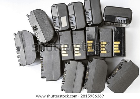 Multiple assorted black digital camera rechargeable lithium-ion battery packs with gold electrical contact pins and connector slots scattered on a clean white background photographed from above showing a variety of sizes and shapes