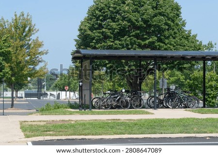 Multiple bicycles parked under a modern covered bike shelter, adjacent to an asphalt parking lot and paved sidewalk, surrounded by lush green trees and bright blue sky, showcasing sustainable urban transportation.