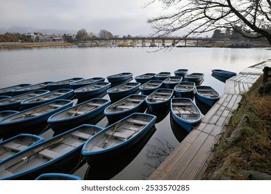 Multiple blue rowboats are docked by a wooden pier on a calm lake with a distant view of a small town and mountains. - Powered by Shutterstock