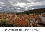 Multiple brick roofs comprising of the Prague Castle Complex and a steep slope filled with vegetation under a thick, cloudy sky are seen
