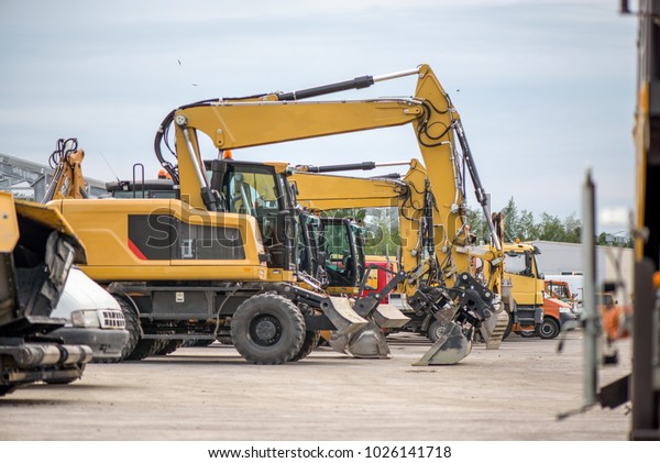 Multiple cars, excavators,\
trucks, loaders, concrete mixers and construction machinery in\
large parking lot in industrial territory, next to concrete and\
asphalt factory
