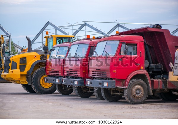 Multiple cars, trucks, loaders, concrete\
mixers and construction machinery in large parking lot in\
industrial territory, next to concrete and asphalt\
factory