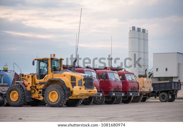 Multiple cars, trucks, loaders, concrete\
mixers and construction machinery in large parking lot in\
industrial territory, next to concrete and asphalt\
factory