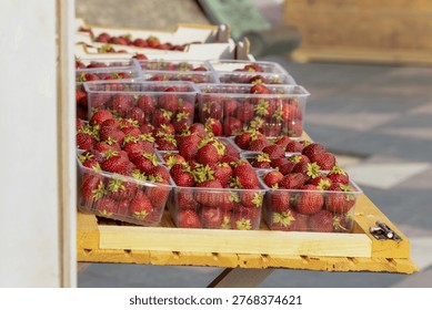 Multiple clear plastic containers filled with ripe red strawberries are arranged on a wooden surface in bright sunlight, creating a vibrant outdoor market presentation with rich color and texture cont