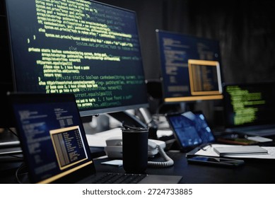 Multiple computer monitors and laptops displaying programming code on screens in dark office environment, showing close up of modern workspace with various electronic devices and coffee cup