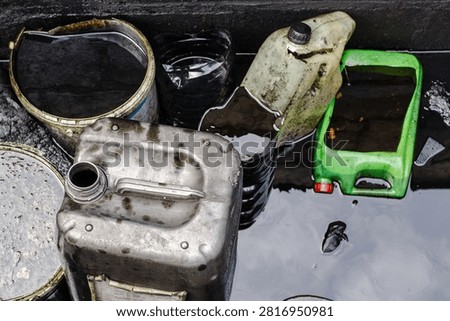 Multiple containers, including metal and plastic jerry cans, are partially submerged in water, highlighting industrial waste and debris in an outdoor setting