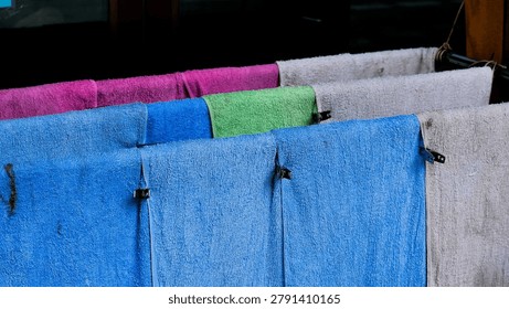 Multiple damp terry cloth towels in blue, pink, magenta, green, and white hang side by side on a clothesline secured with black metal clips, shot close-up against a dark background, conveying a sense of everyday domestic life.