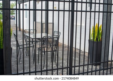 Multiple Empty Black Metal Tables And Chairs On A Cafe Sidewalk Patio Of A Restaurant. The Exterior Brick Wall And Background Are White And There's A Black Fence And Gate With Potted Flowers In Front.